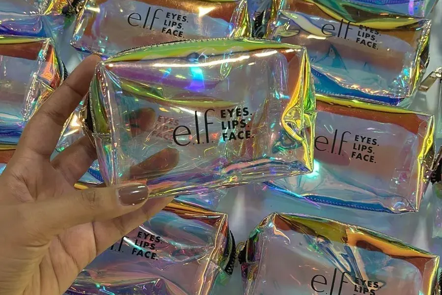 Production run of clear iridescent ELF COSMETICS cosmetic pouches, with a hand-held pouch showing the black elf wordmark and EYES. LIPS. FACE. branding